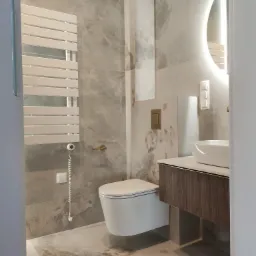 Wykończona łazienka z toaletą typu Geberit, umywalką nablatową na szafce z ciemnego drewna i podświetlanym lustrem. Na ścianach i podłodze płytki imitujące marmur.