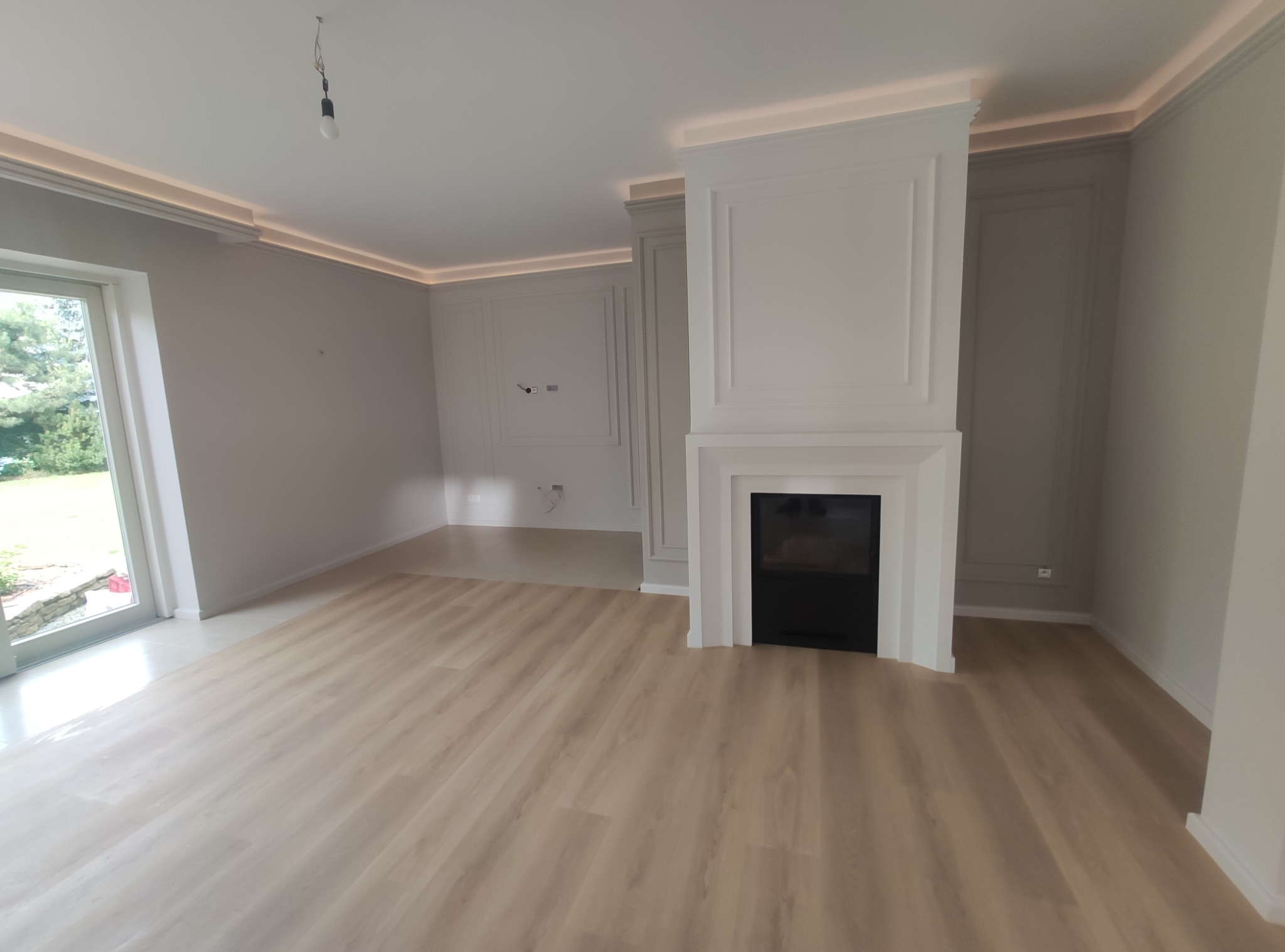 Jasne panele podłogowe w nowo wykończonym salonie z kominkiem i sztukaterią, oświetlenie LED na suficie.