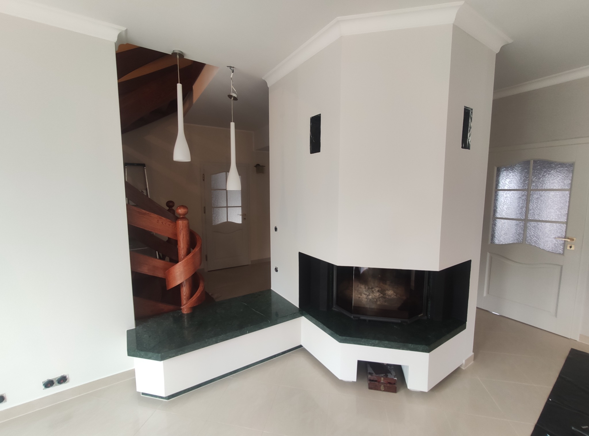 Nowoczesny salon z białym kominkiem obłożonym zielonym marmurem, spiralnymi drewnianymi schodami i dwoma wiszącymi lampami o stożkowym kształcie.