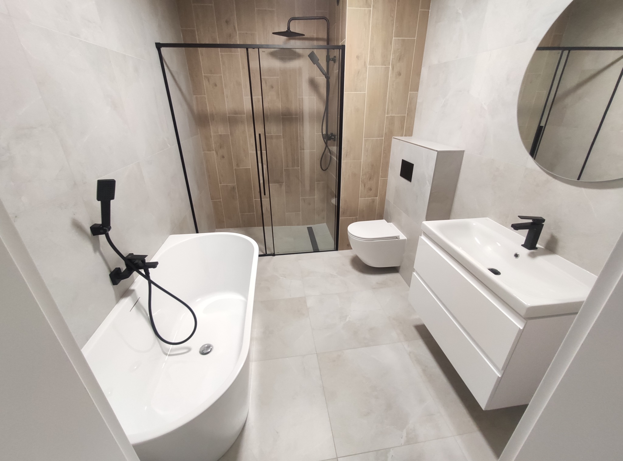 Nowoczesna łazienka z wanną wolnostojącą, kabiną prysznicową typu walk-in z czarnymi ramami, umywalką nablatową z szafką i okrągłym lustrem, wykończona płytkami w odcieniach beżu i szarości.