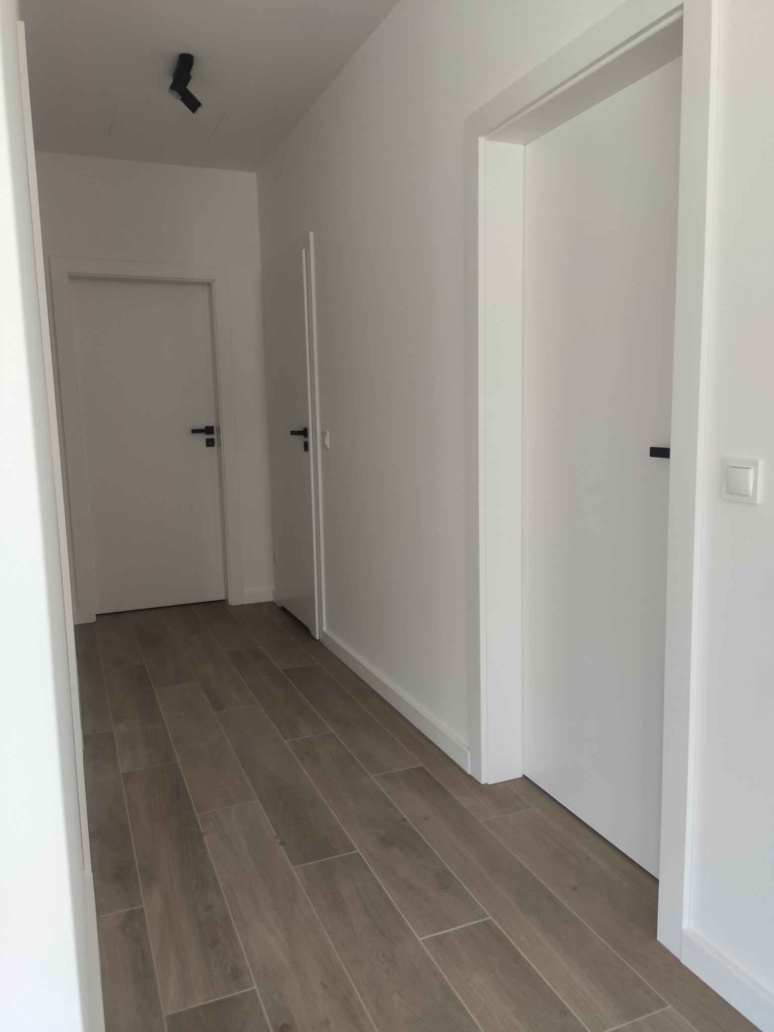 Korytarz w jasnych barwach z drewnopodobną podłogą, trzema białymi drzwiami z czarnymi klamkami i punktowym oświetleniem sufitowym.