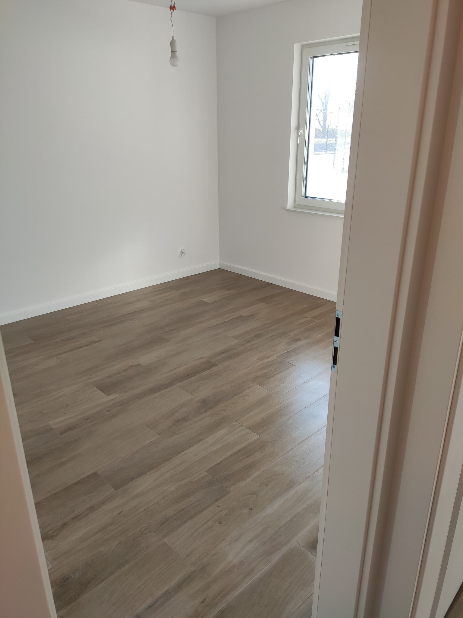 Pusty pokój z ułożonymi panelami podłogowymi w odcieniach brązu, widok zza uchylonych drzwi, białe ściany, okno z widokiem na zewnątrz, wisząca żarówka bez klosza.