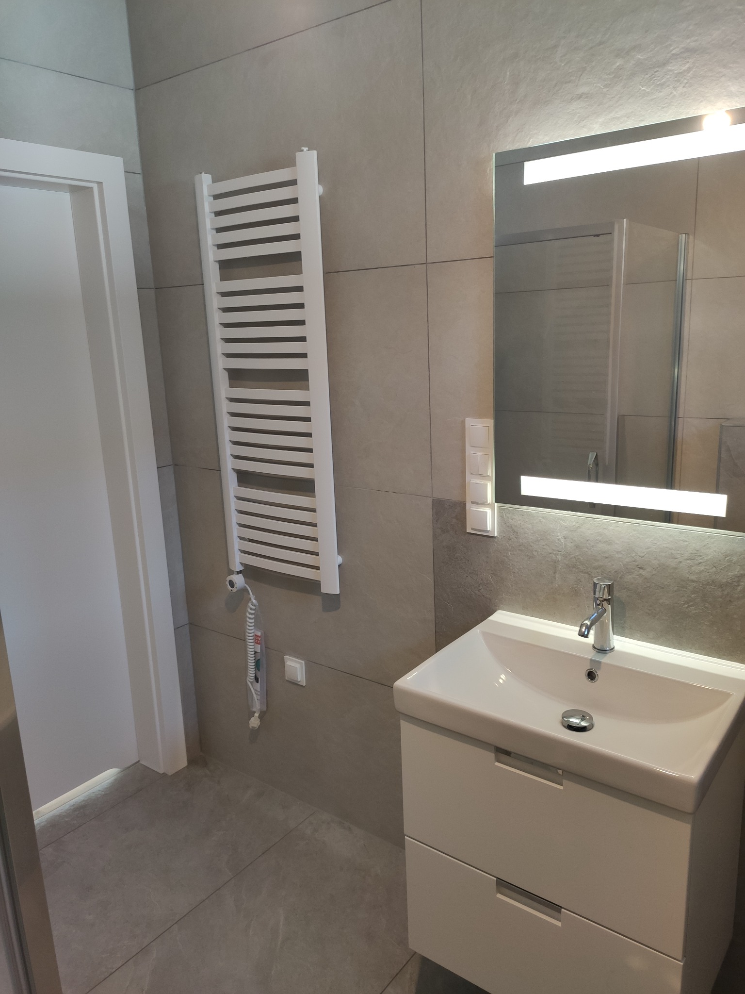 Nowoczesna łazienka z szarymi płytkami, białym grzejnikiem drabinkowym, umywalką z szafką i lustrem podświetlanym LED.