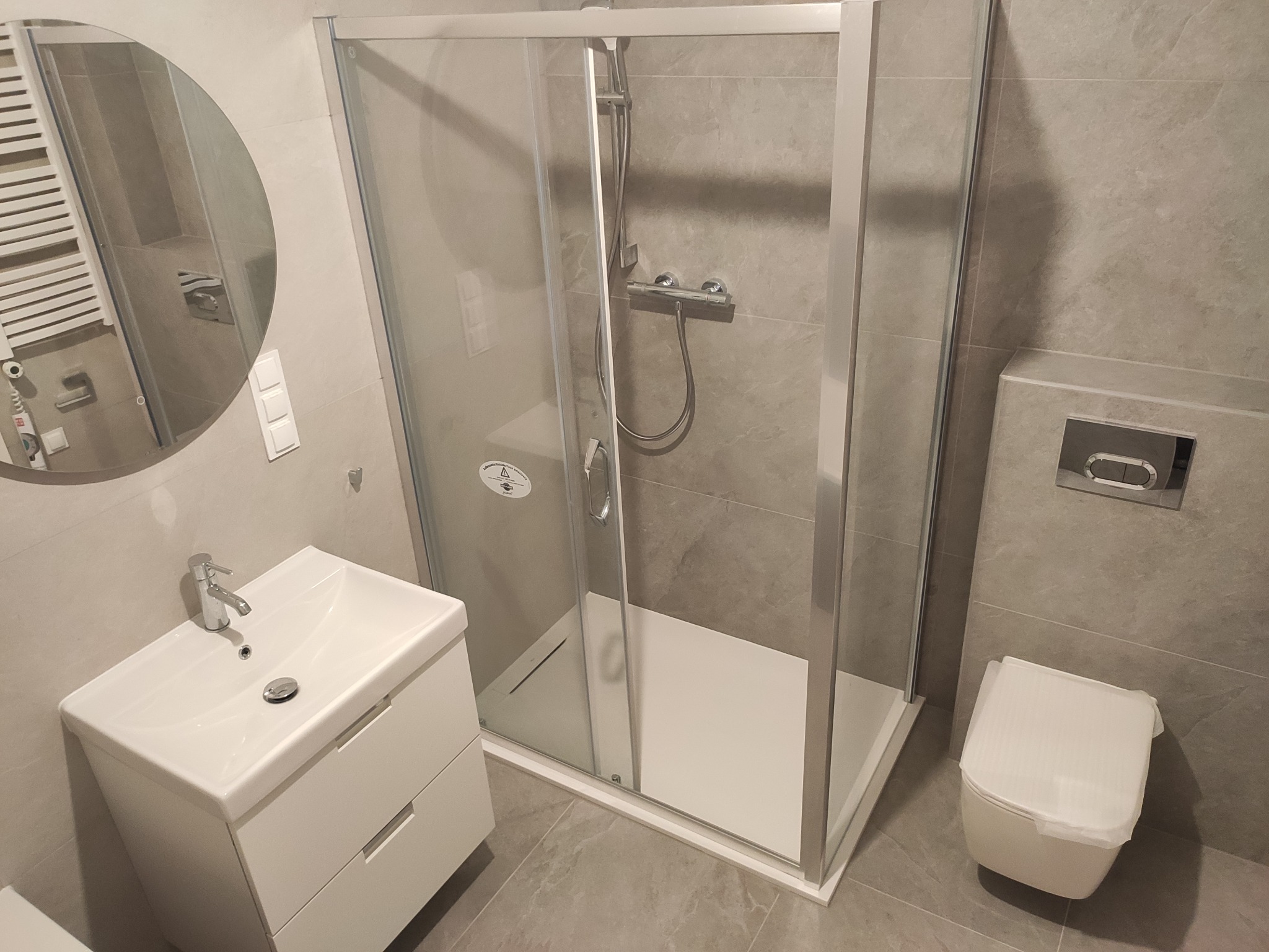 Nowoczesna łazienka w szarościach z kabiną prysznicową, umywalką z szafką i podwieszaną toaletą, okrągłe lustro odbija grzejnik drabinkowy.