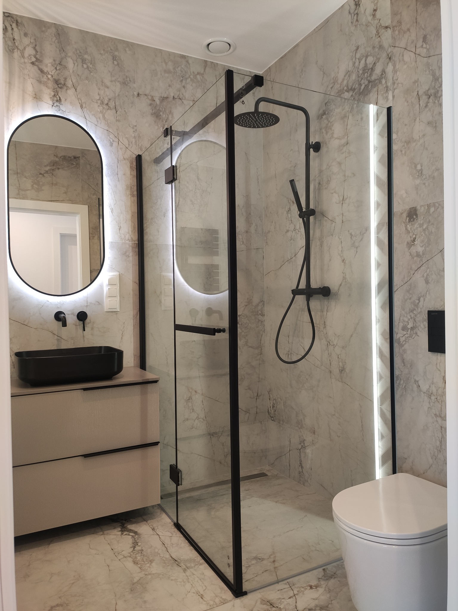 Nowoczesna łazienka z kabiną prysznicową typu walk-in, umywalką nablatową i podświetlanym lustrem. Marmurowe płytki na ścianach i podłodze.