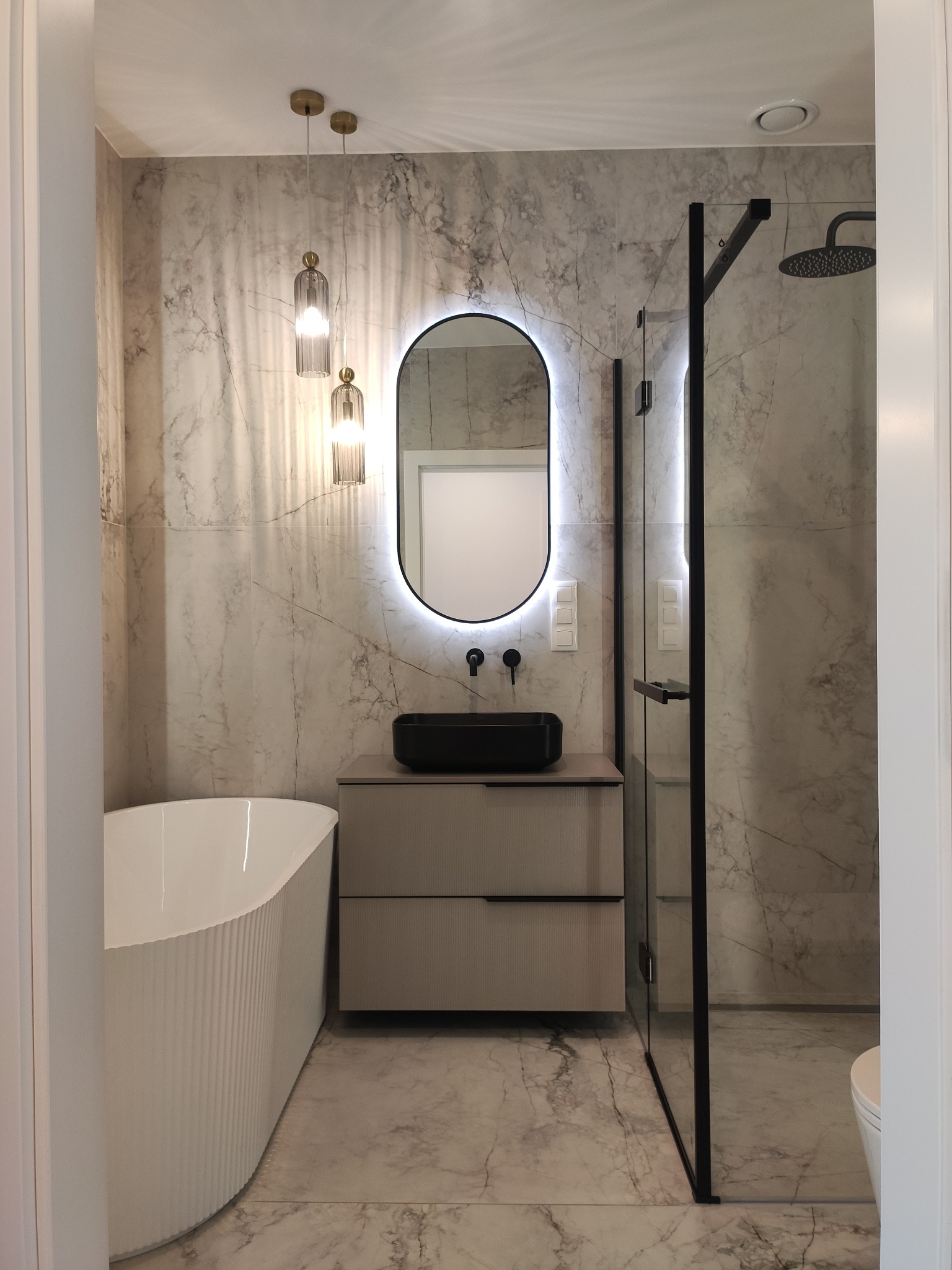 Elegancka łazienka z wanną, czarną umywalką na szafce i kabiną prysznicową. Ściany i podłoga wyłożone marmurowymi płytkami. Oświetlenie punktowe i lustro z podświetleniem LED.