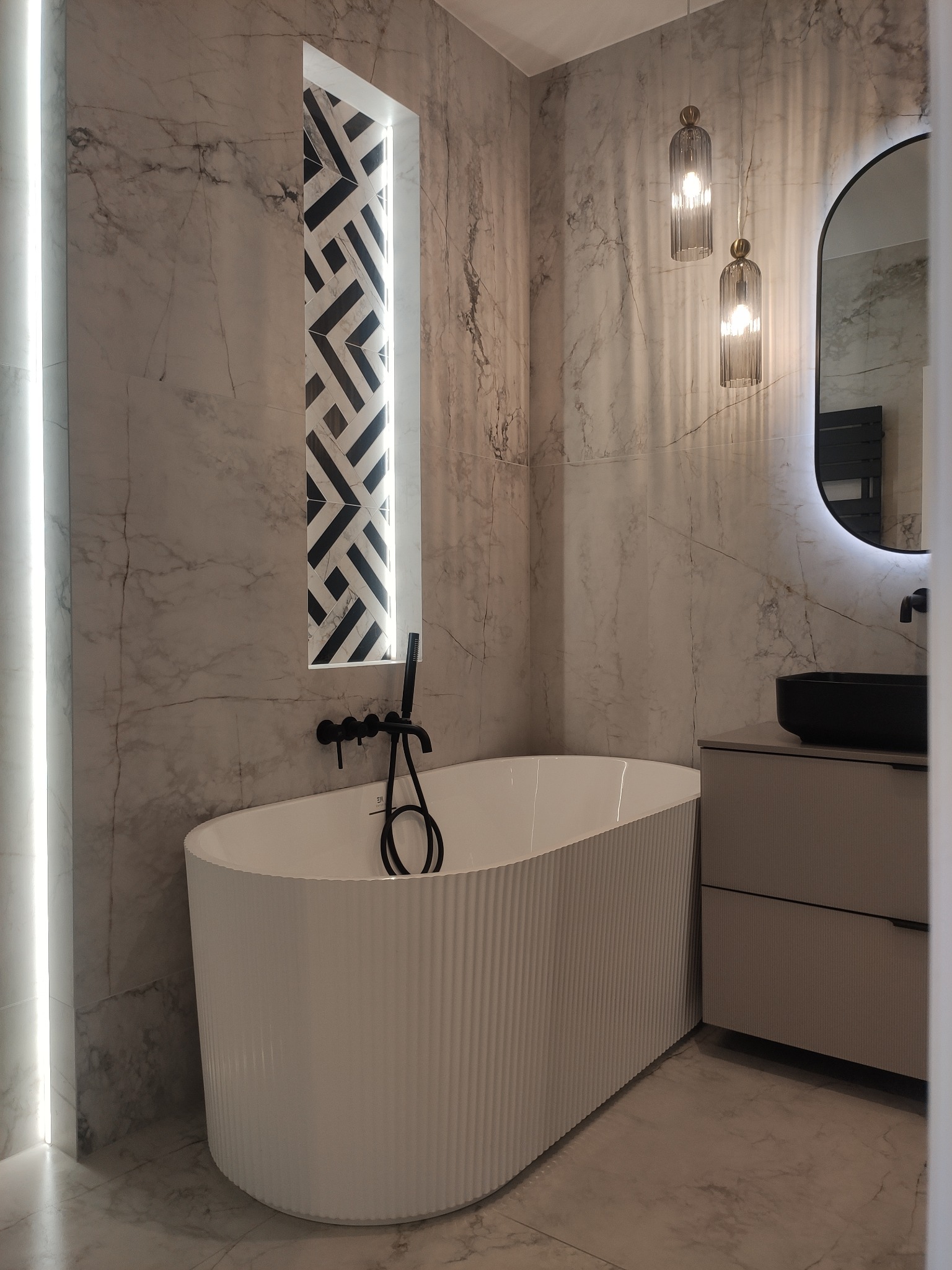 Elegancka łazienka z wanną z żebrowaną obudową, marmurowymi płytkami i czarną armaturą. Wnęka z geometrycznym wzorem i podświetleniem LED.