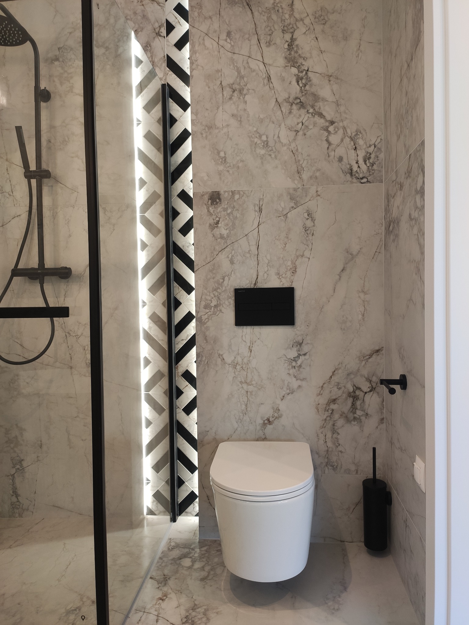 Wykończona łazienka w marmurowej tonacji z kabiną prysznicową typu walk-in, podświetlanym panelem dekoracyjnym i wiszącą toaletą.