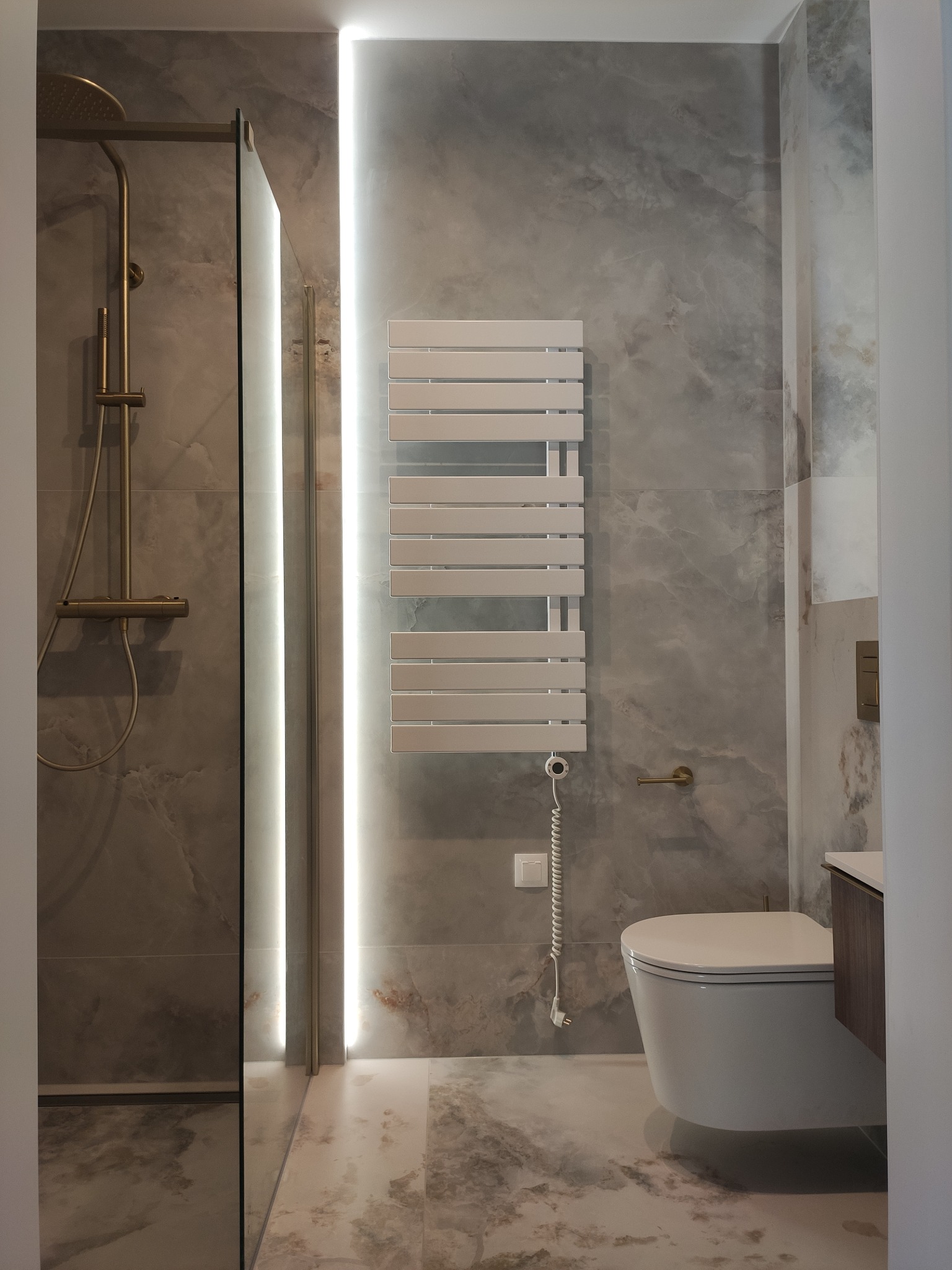 Elegancka łazienka z marmurowymi płytkami, złotą armaturą i podświetlanym grzejnikiem ręcznikowym. Nowoczesny design z dbałością o detale.