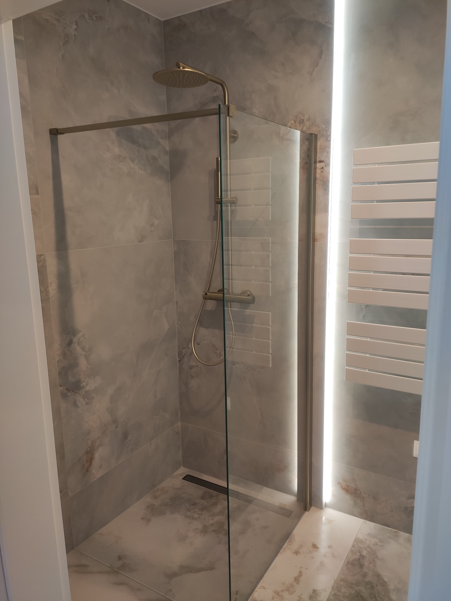 Nowoczesna kabina prysznicowa z deszczownicą w łazience wykończonej płytkami imitującymi marmur, z podświetlanym grzejnikiem drabinkowym.