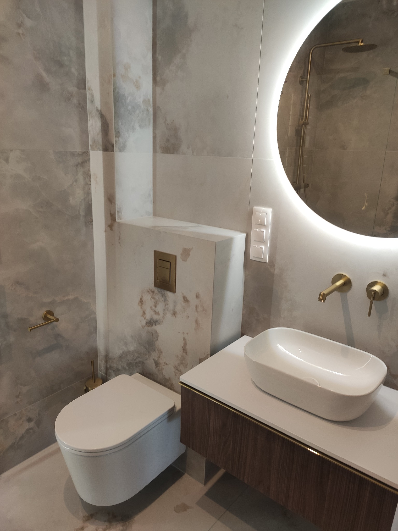 Nowoczesna łazienka z umywalką nablatową, podświetlanym lustrem, toaletą podwieszaną i płytkami imitującymi marmur, złote akcenty armatury.