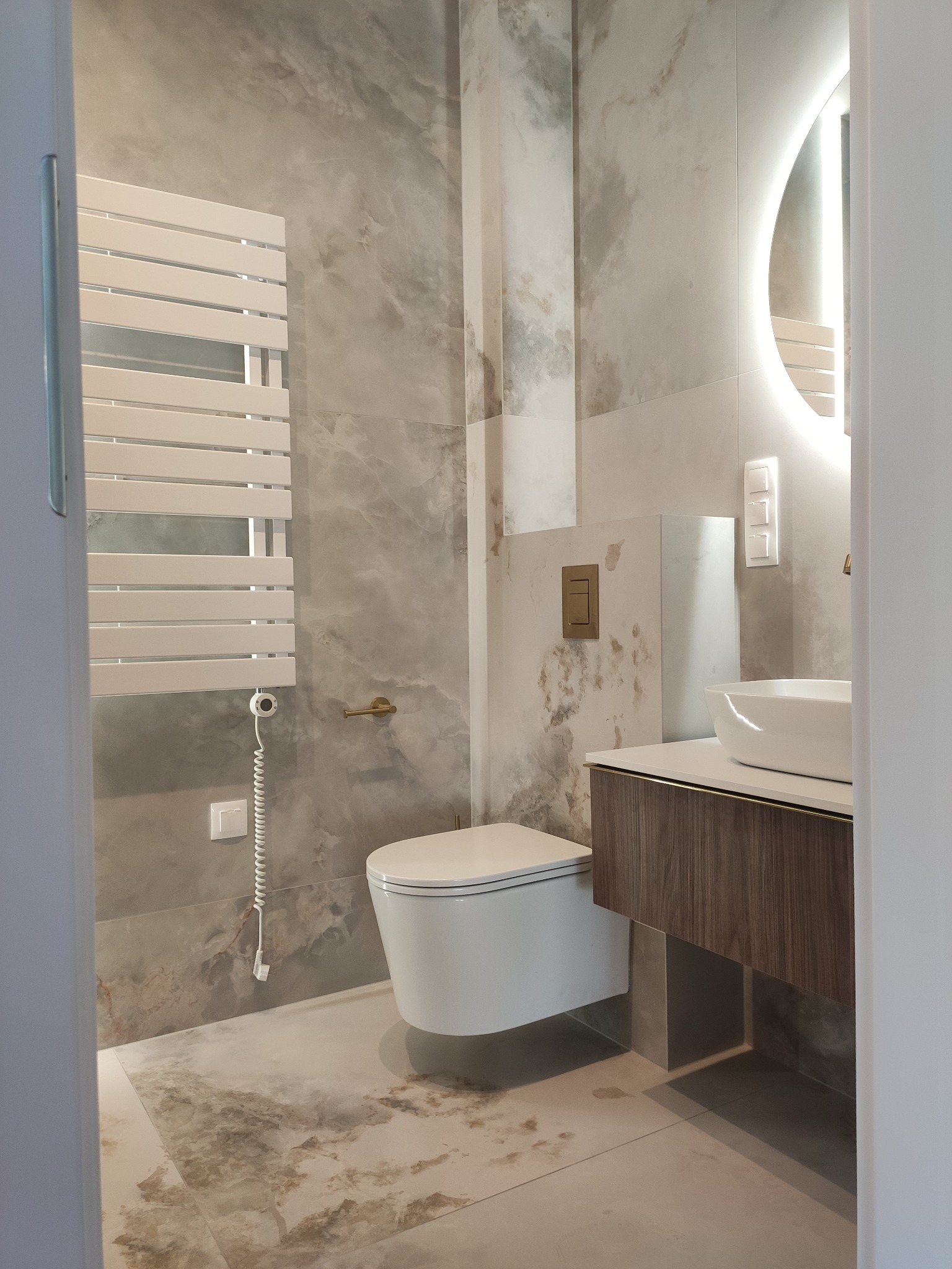 Wykończona łazienka z toaletą typu Geberit, umywalką nablatową na szafce z ciemnego drewna i podświetlanym lustrem. Na ścianach i podłodze płytki imitujące marmur.