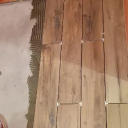 Układanie drewnopodobnych paneli podłogowych z użyciem krzyżyków dystansowych, widoczna warstwa kleju na podłożu i pomarańczowa ściana w tle.