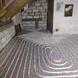 Rozłożone rury ogrzewania podłogowego Reflex Thermosystem na izolacji podposadzkowej w surowym wnętrzu z bloków betonowych i drewnianą więźbą dachową.