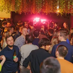 Tłum ludzi w barze z neonowym napisem 'All you need is love' w tle, dekoracje z roślin zwisających z sufitu, mężczyzna z piwem na pierwszym planie.