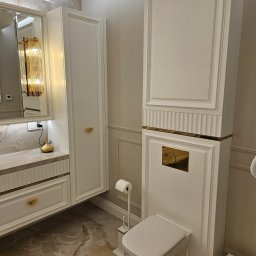 StarBud - Elegancka łazienka w odcieniach bieli i złota: wisząca toaleta, szafki z frezowanymi frontami i marmurowa podłoga. Lampa w stylu glamour odbija się w lustrze.