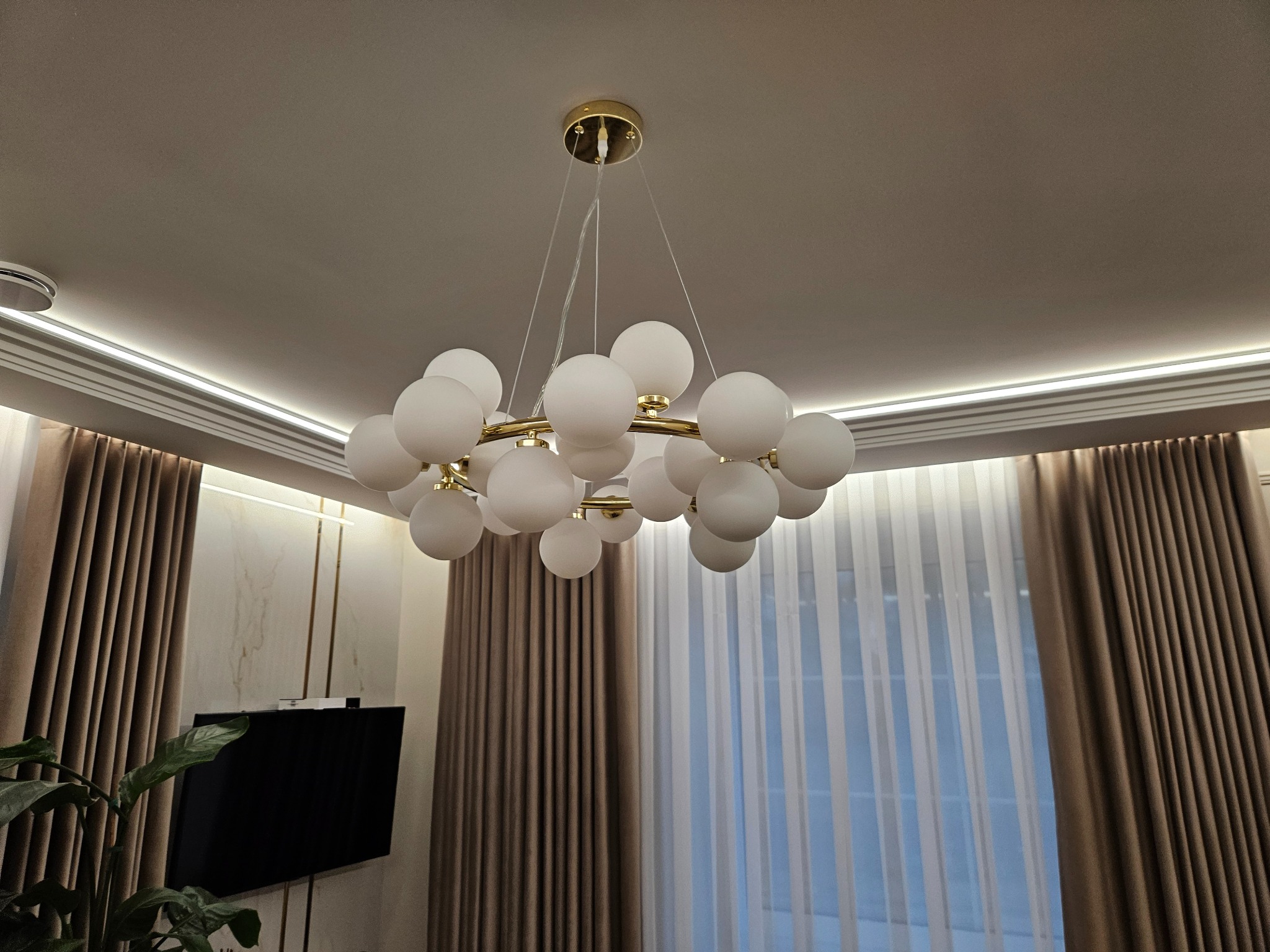 Nowoczesny żyrandol z białymi kulami i złotym okręgiem, zawieszony w eleganckim salonie z zasłonami i podświetleniem sufitowym LED.