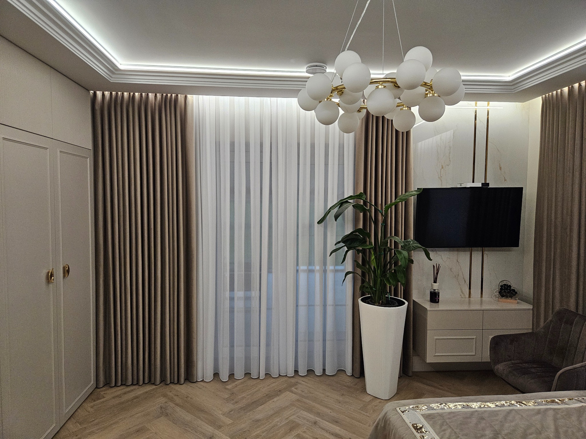 Eleganckie wnętrze: beżowe zasłony, biały tiul, parkiet w jodełkę, designerska lampa i telewizor na marmurowej ścianie z złotymi akcentami.