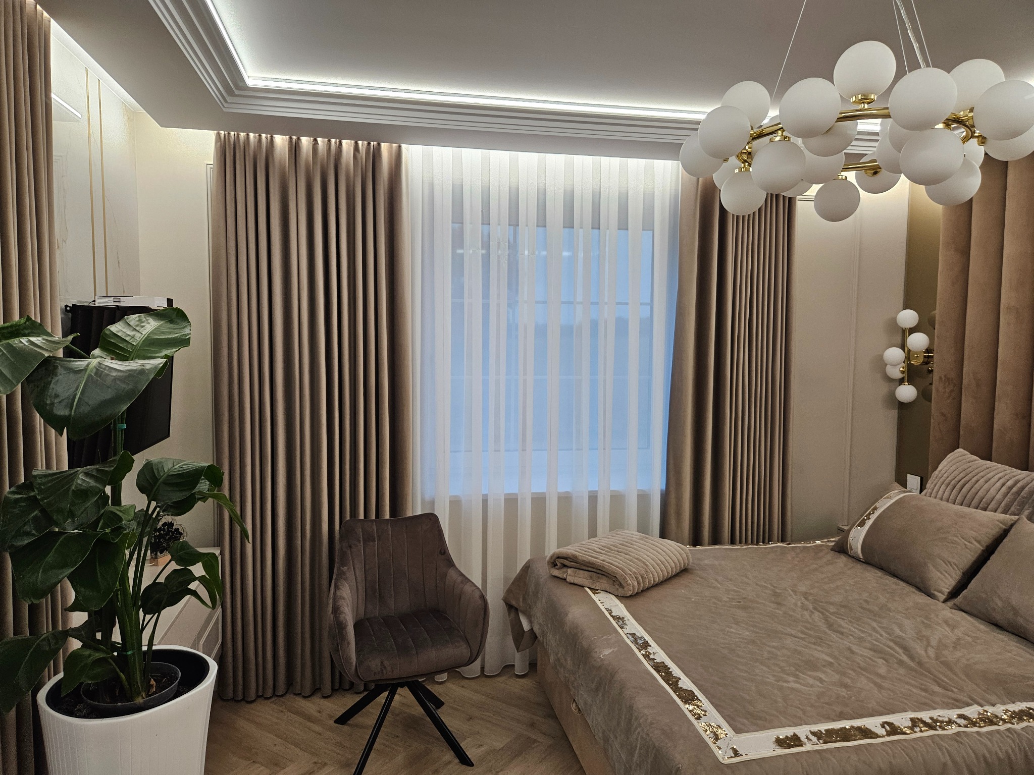 Elegancka sypialnia w beżach: miękkie zasłony, designerska lampa, wygodne łóżko z dekoracyjnym wykończeniem i fotel na tle parkietu w jodełkę.