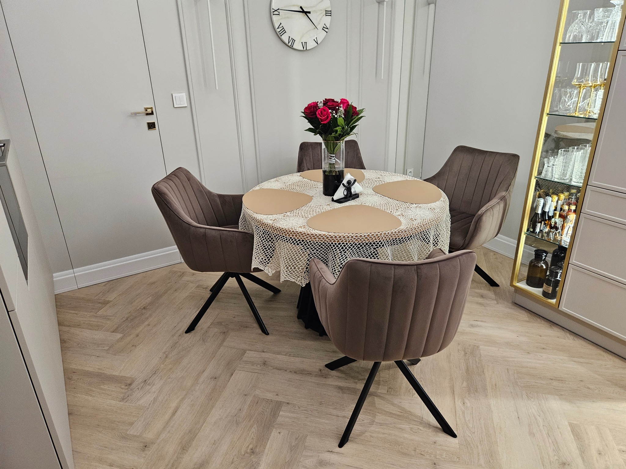 Elegancka jadalnia z parkietem w jodełkę, okrągłym stołem z bukietem róż i krzesłami w kolorze taupe. W tle białe ściany z dekoracyjnymi listwami i przeszklona witryna.