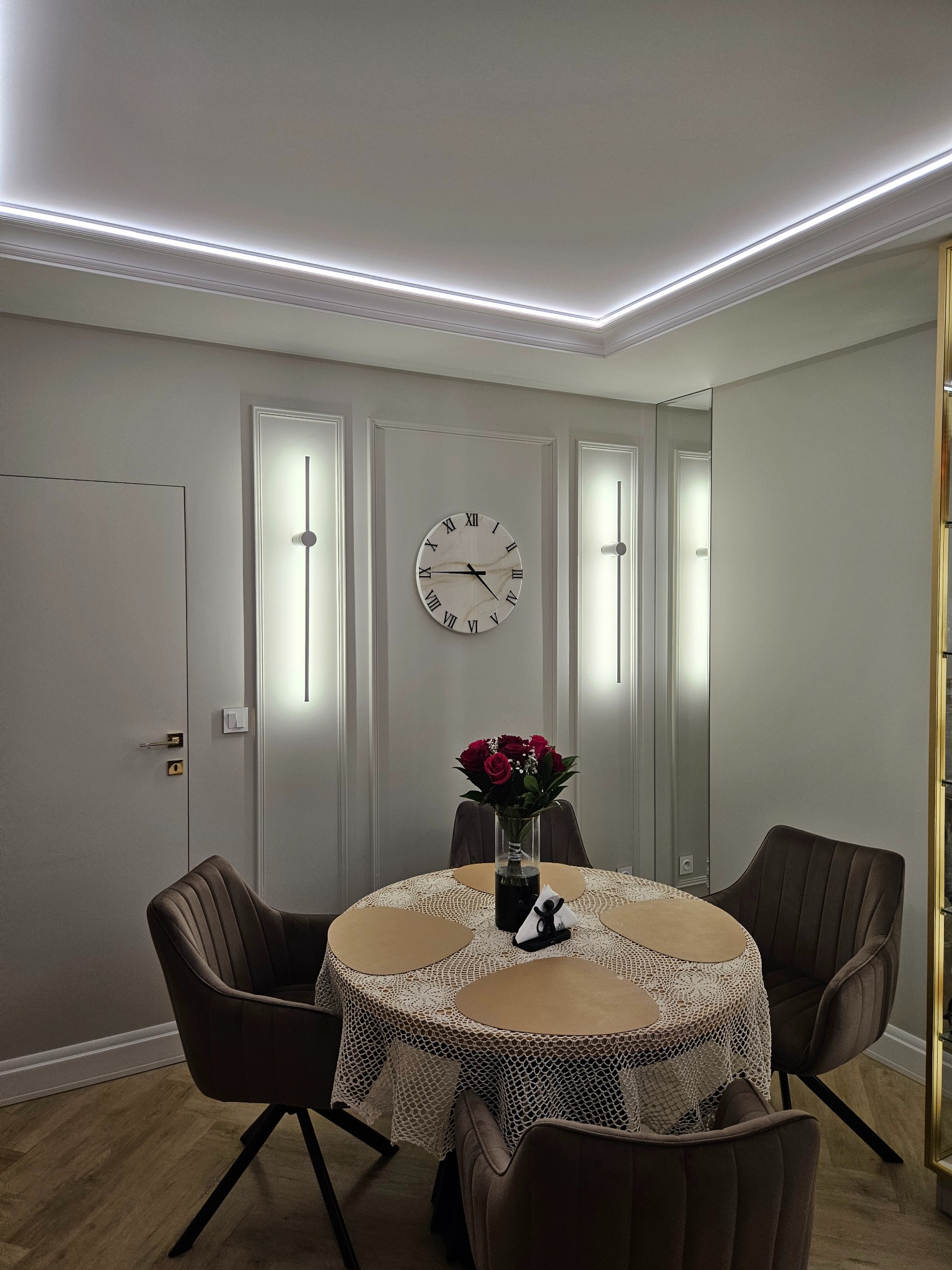 Elegancka jadalnia w stonowanej kolorystyce: okrągły stół z koronkowym obrusem, welurowe krzesła, zegar na ścianie i subtelne oświetlenie LED.