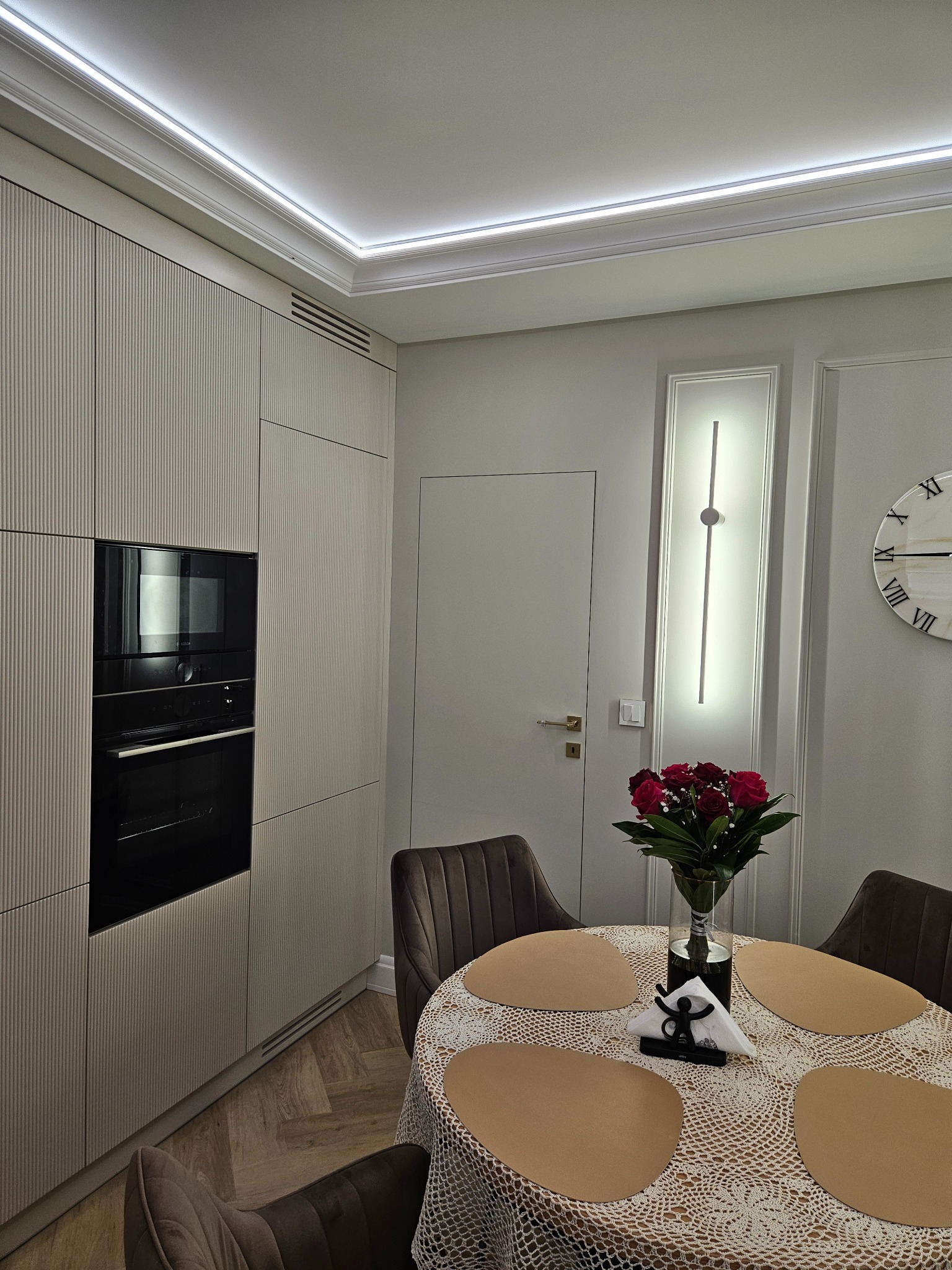 Elegancka kuchnia w odcieniach beżu z frezowanymi frontami, oświetleniem LED podszafkowym i nowoczesnym piekarnikiem. Na stole bukiet czerwonych róż.