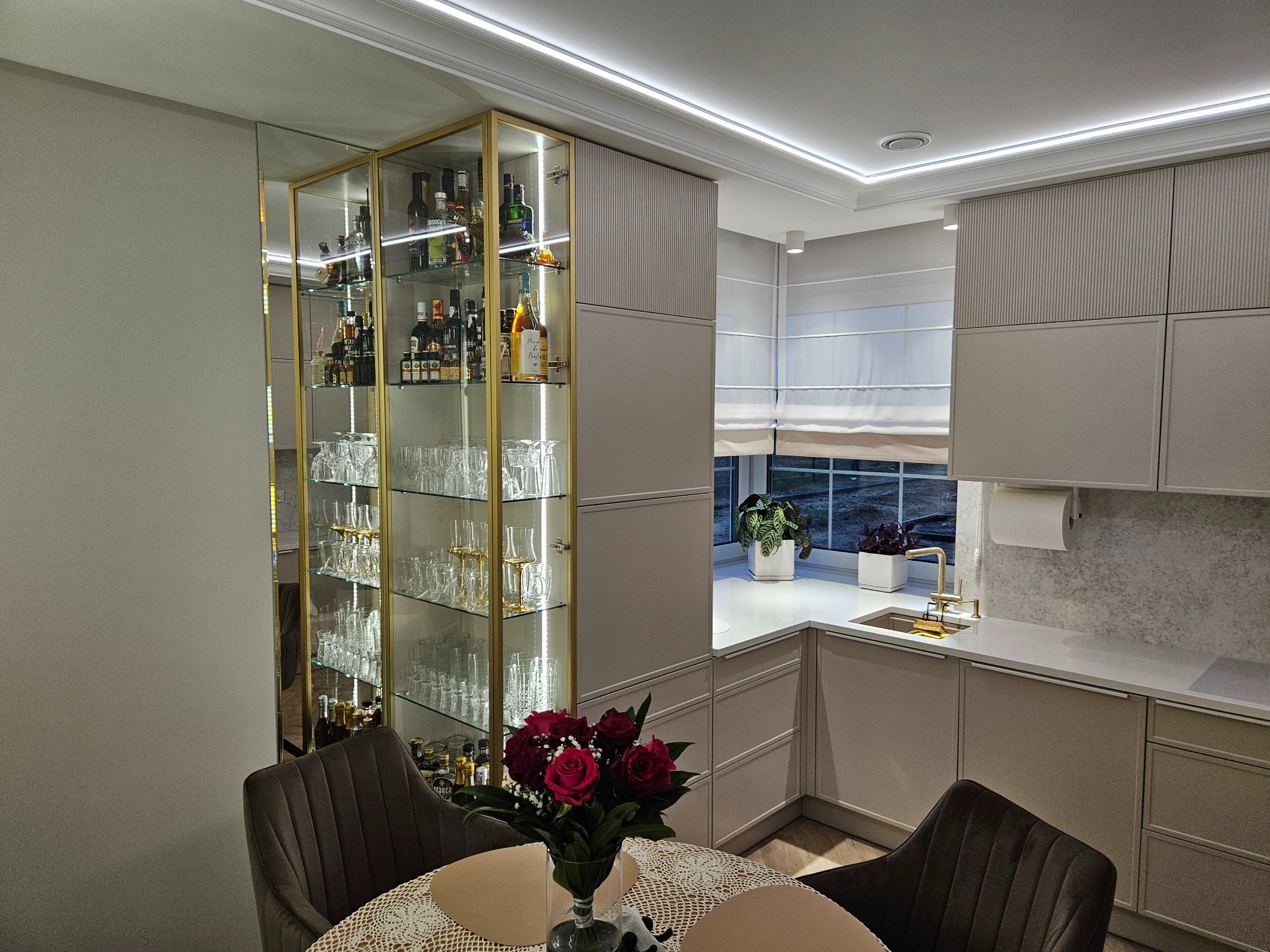 Eleganckie wykończenie wnętrza kuchni z przeszkloną witryną na alkohol, blatem z rośliną i bukietem czerwonych róż na stole. Oświetlenie LED podkreśla nowoczesny design.