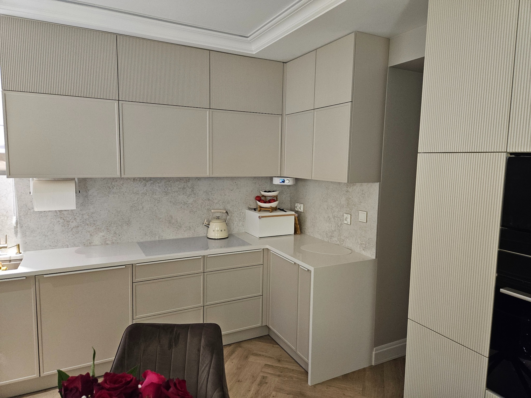 Beżowa kuchnia z frezowanymi frontami szafek górnych, jasnym blatem i drewnianą podłogą w jodełkę. Na blacie czajnik Smeg i pojemnik na pieczywo.