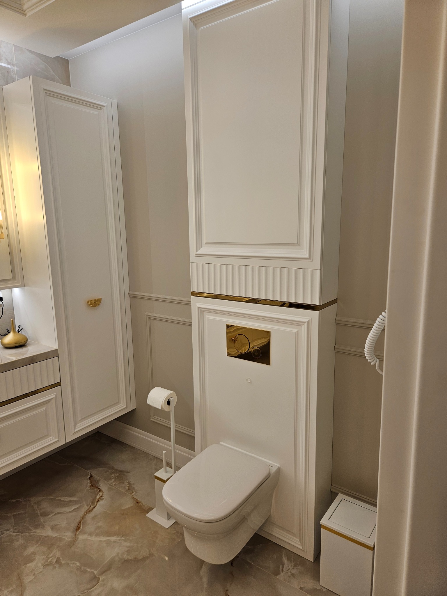 Elegancka łazienka w odcieniach bieli i złota: zabudowana toaleta z szafką, marmurowe płytki podłogowe, detale dekoracyjne. Nowoczesny design z luksusowymi akcentami.
