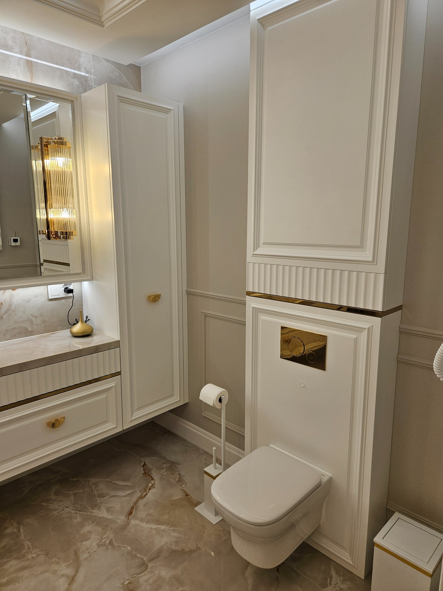 Elegancka łazienka w odcieniach bieli i złota: wisząca toaleta, szafki z frezowanymi frontami i marmurowa podłoga. Lampa w stylu glamour odbija się w lustrze.