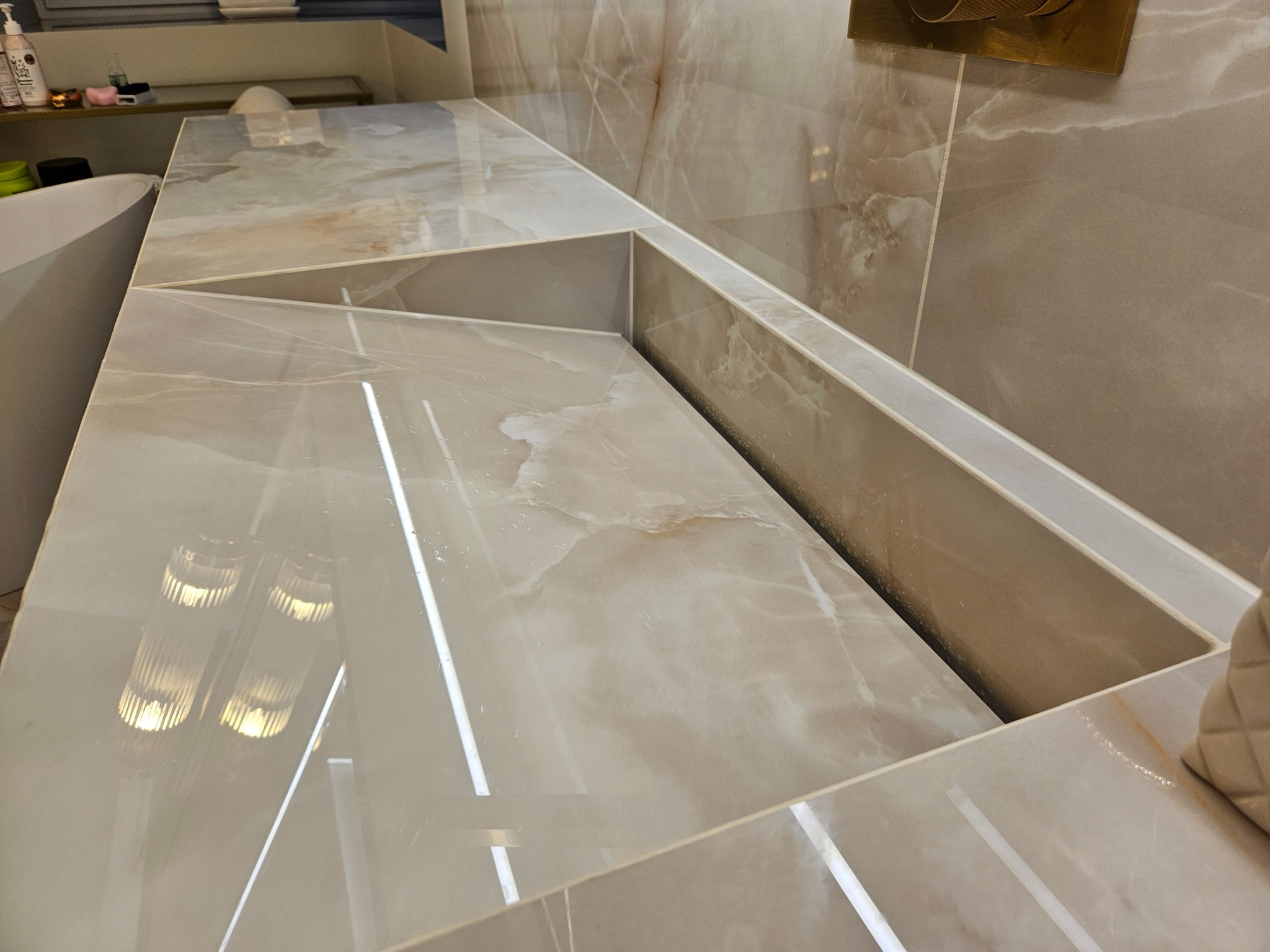 Beżowa, marmurkowa umywalka w łazience, z widocznymi refleksami światła. Umywalka zintegrowana z blatem, wykonana z płytek. Minimalistyczny design, eleganckie wykończenie.