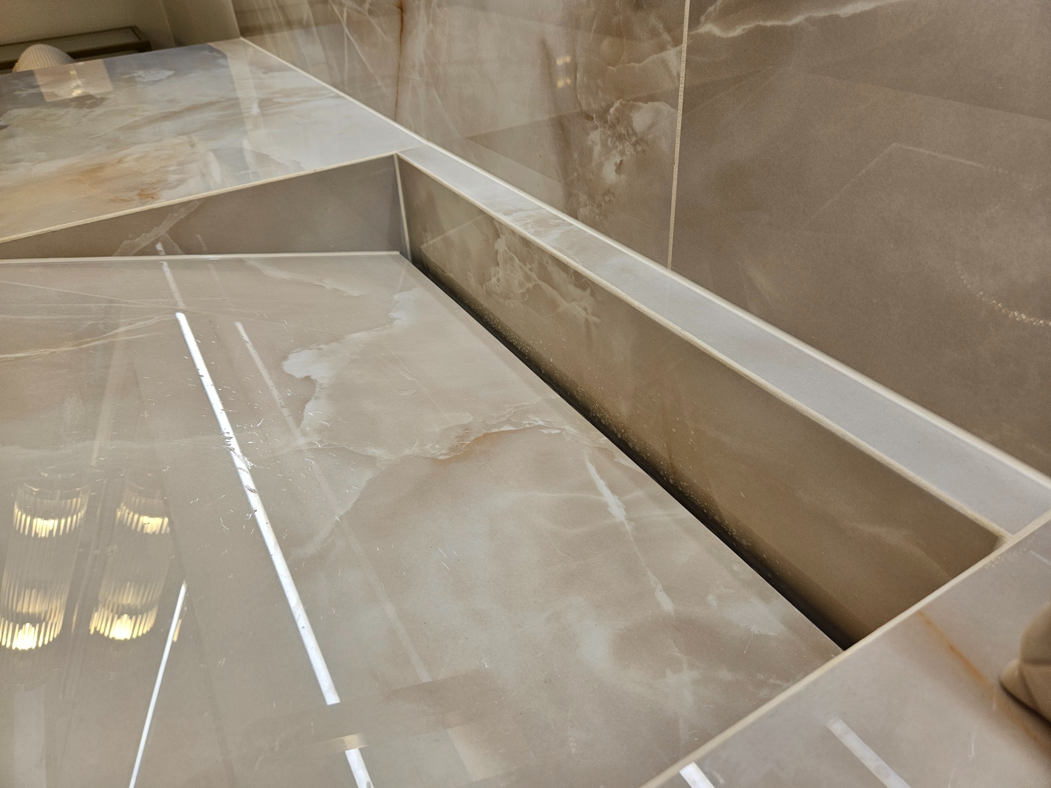 Beżowe, polerowane płytki imitujące marmur, ułożone w geometryczny wzór na ścianie i blacie. Widoczne fugi i odbicie światła, podkreślające gładką powierzchnię.