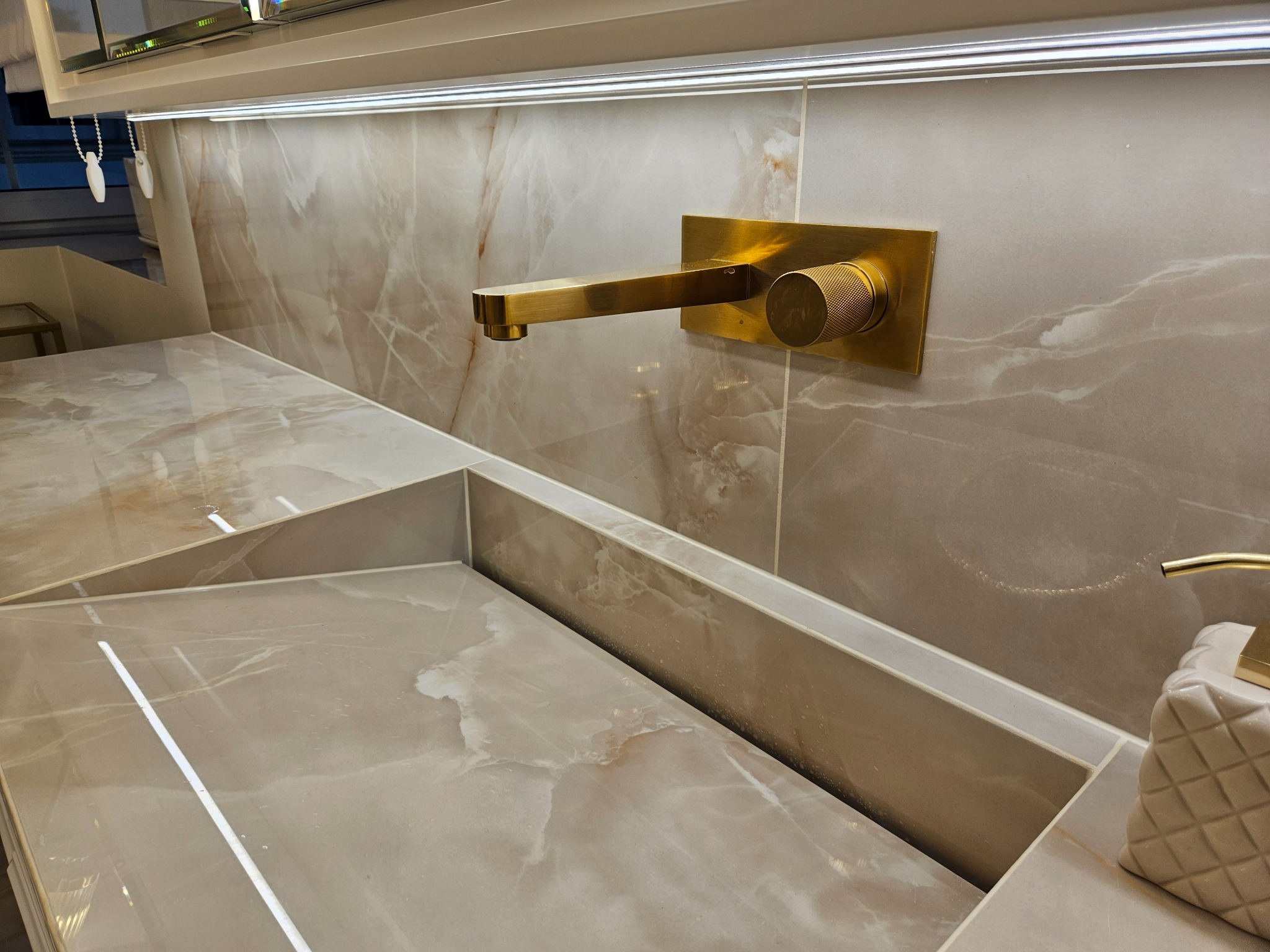 Beżowa umywalka z marmurowym wzorem i złotą armaturą ścienną. Minimalistyczny design, oświetlenie LED podkreśla elegancję wykończenia.