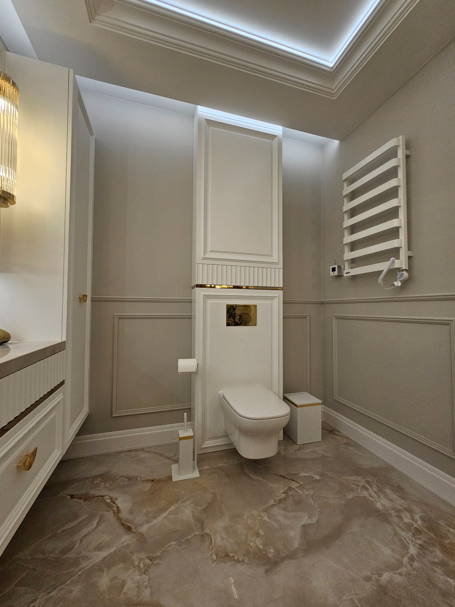 Elegancka łazienka w odcieniach beżu i bieli z marmurową podłogą. Uwagę zwraca zabudowana toaleta z elementami dekoracyjnymi w kolorze złota i grzejnik drabinkowy.