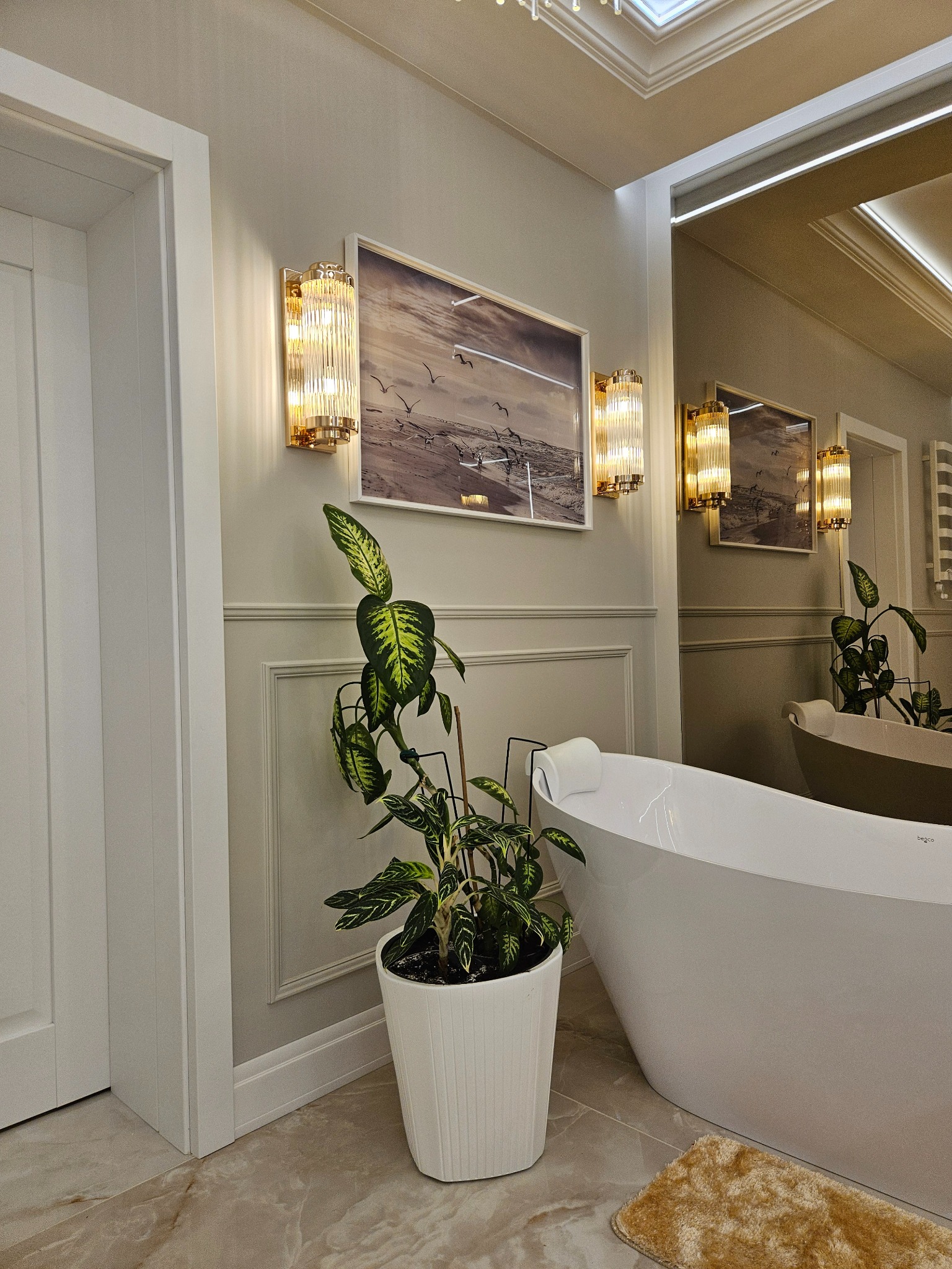 Elegancka łazienka z wolnostojącą wanną, oświetleniem ściennym i dekoracyjną rośliną w białej doniczce. Na ścianie obraz z motywem ptaków.