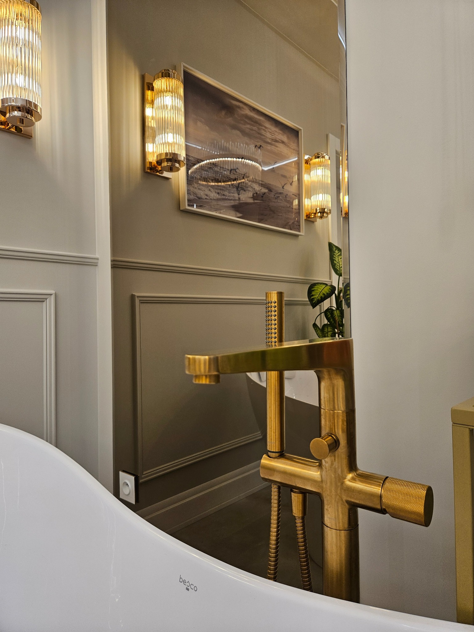 Elegancka bateria wannowa w kolorze złotym, na tle stylowej łazienki z panelami ściennymi i oświetleniem. Widoczny fragment wanny i dekoracje.