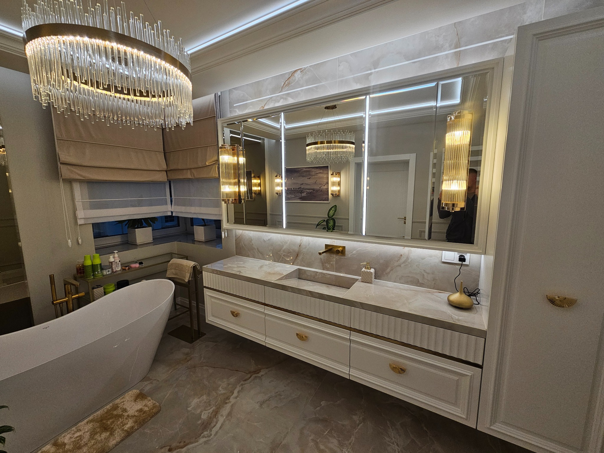 Elegancka łazienka w jasnych barwach z marmurową posadzką i blatem. Uwagę przykuwa designerski żyrandol i duże lustro z oświetleniem LED. Widoczna wanna wolnostojąca.