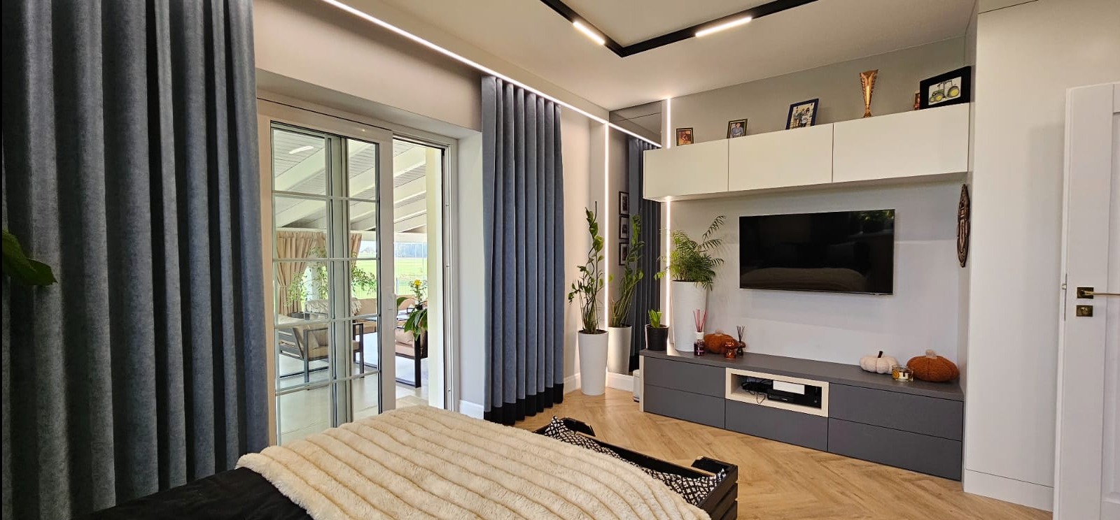 Nowoczesny salon z szarymi zasłonami, telewizorem na ścianie i parkietem w jodełkę. Widok na taras przez przeszklone drzwi. Oświetlenie LED podkreśla minimalistyczny design.