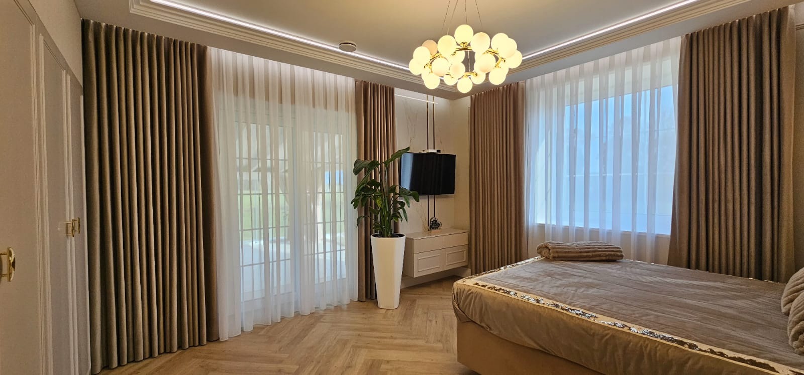 Elegancka sypialnia z beżowymi zasłonami i firanami, drewnianą podłogą w jodełkę, nowoczesną lampą i telewizorem na ścianie. Wnętrze po remoncie, gotowe do zamieszkania.
