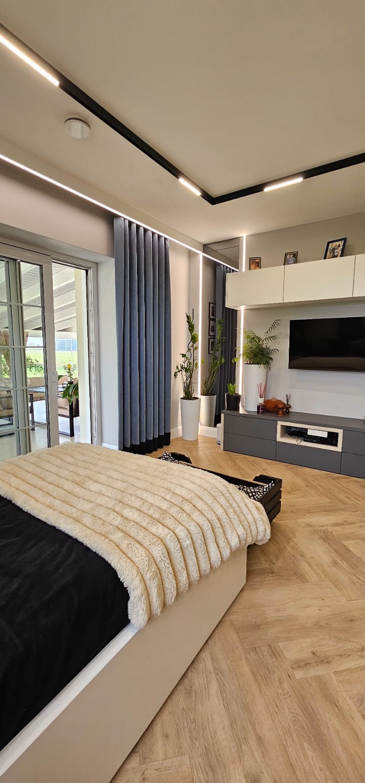 Nowoczesna sypialnia z parkietem w jodełkę, oświetleniem LED na suficie i szarymi zasłonami. Widoczne rośliny doniczkowe i telewizor na ścianie.