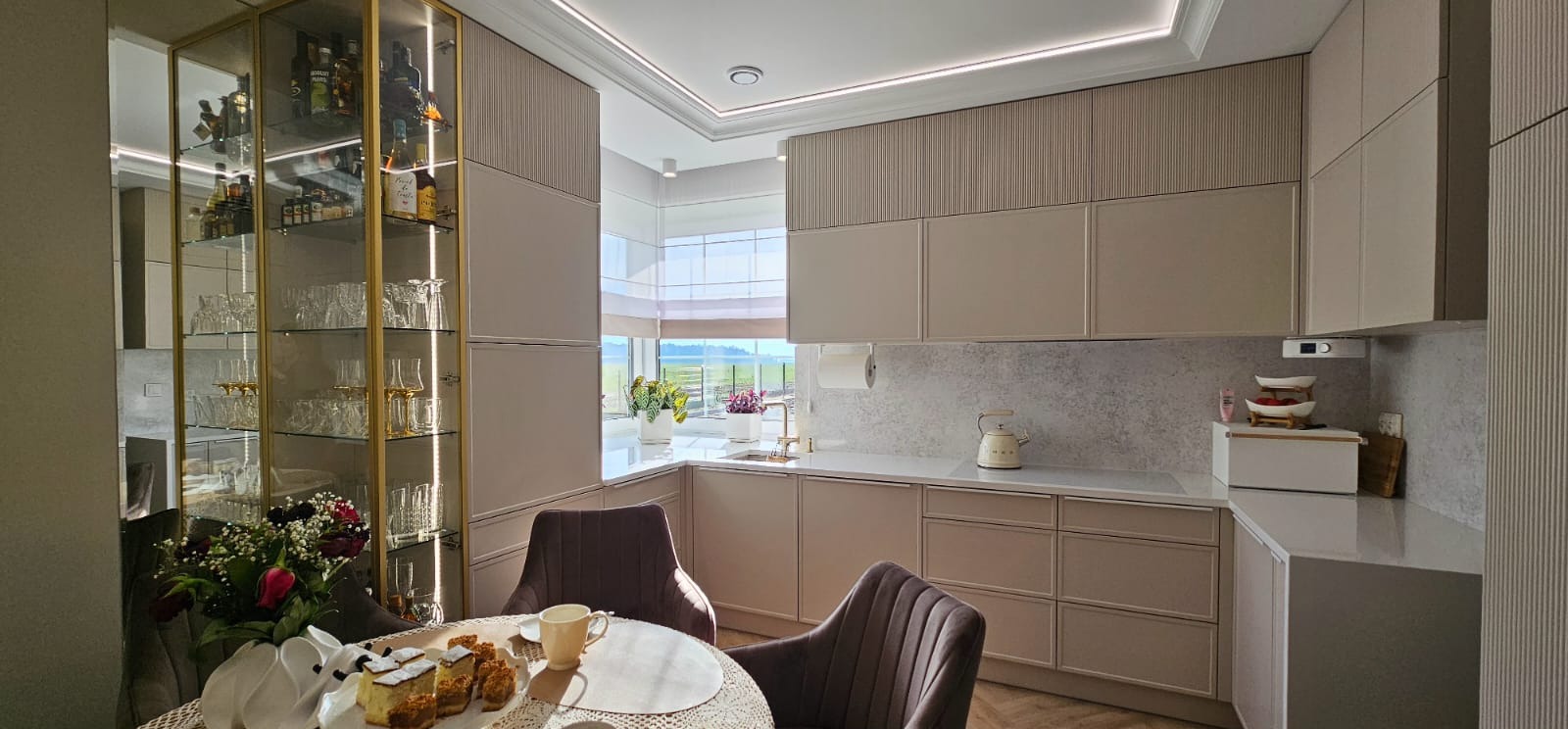 Jasna kuchnia w stylu nowoczesnym z przeszkloną witryną, okrągłym stołem i krzesłami. Widoczne detale wykończenia i oświetlenie LED.