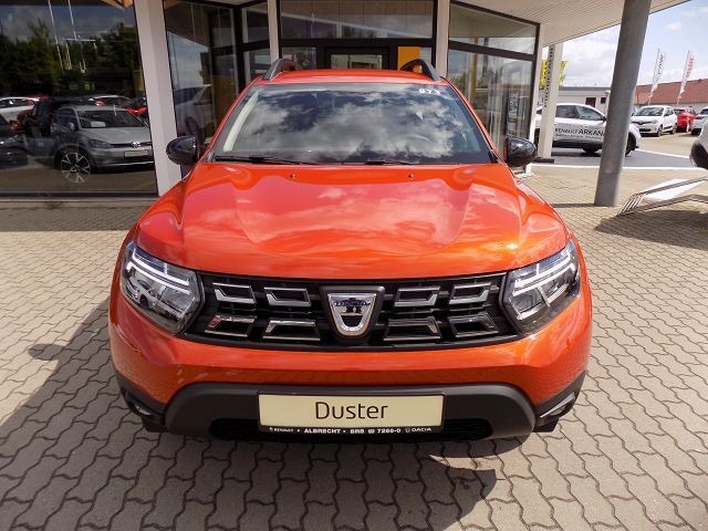 Pomarańczowy samochód Dacia Duster z przodu, zaparkowany na placu dealera samochodowego z widocznymi innymi pojazdami w tle.