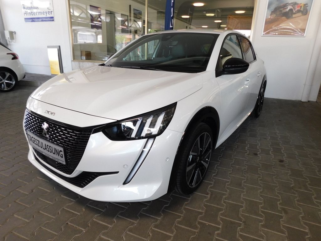 Biały samochód Peugeot 208 z czarnymi felgami na tle salonu samochodowego z widocznym plakatem na ścianie.