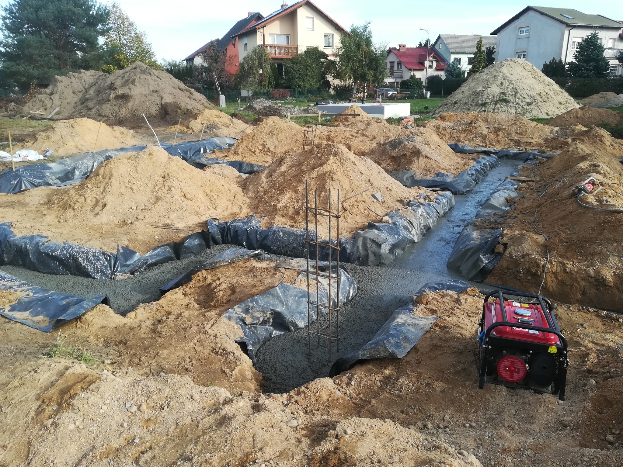 Wylane fundamenty zbrojone stalą, zabezpieczone folią, widoczny generator prądu na placu budowy, w tle domy jednorodzinne i hałdy piasku.
