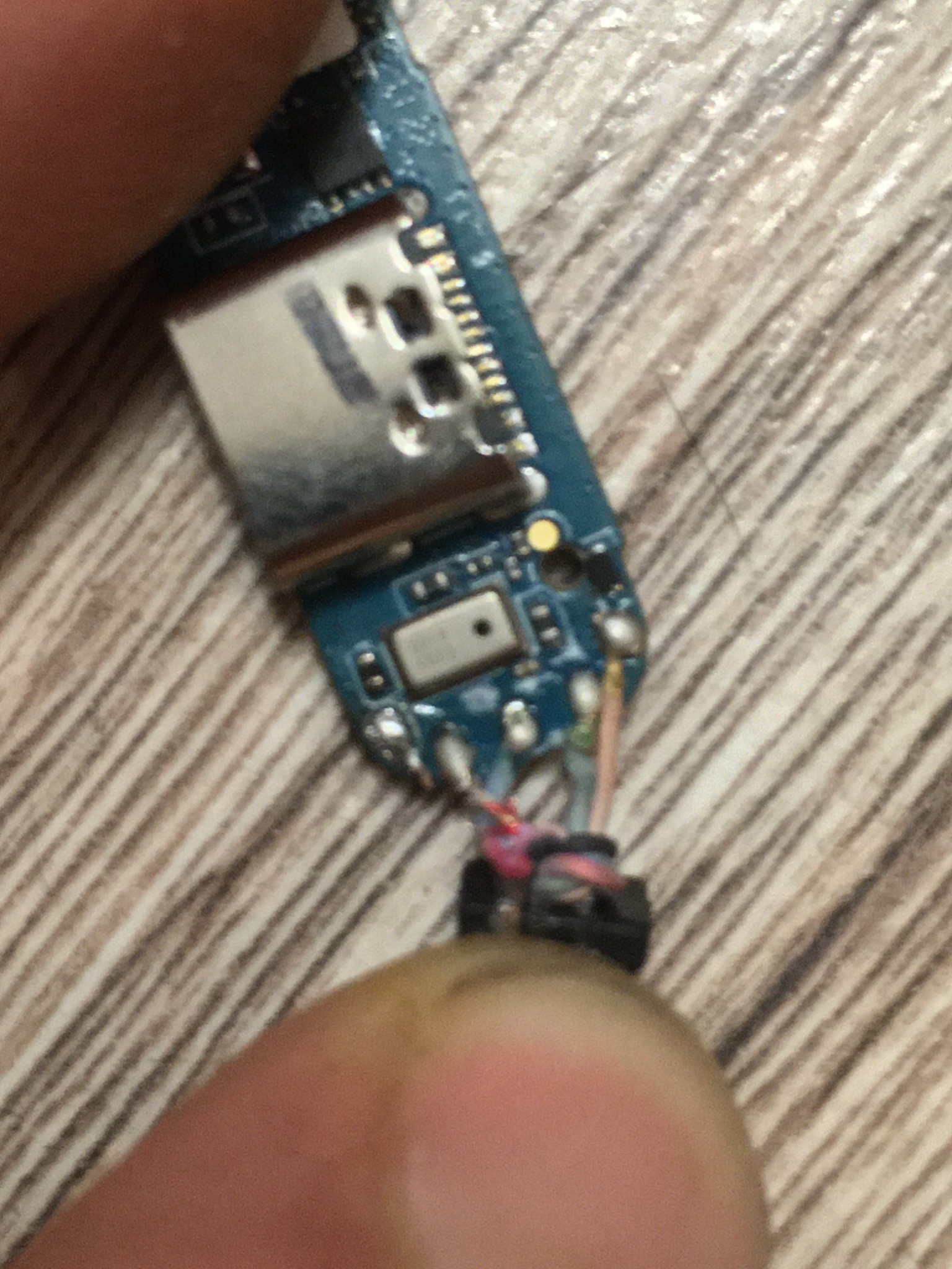 Detalik elektroniki z widocznymi ścieżkami i elementami lutowanymi, trzymany w palcach na tle drewnopodobnej powierzchni.