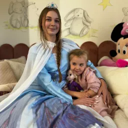 Animator w stroju Elsy z Krainy Lodu obejmuje dziewczynkę trzymającą lalkę Elsy na tle ściany z dziecięcymi rysunkami.