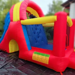 Kolorowy, dmuchany zamek ze zjeżdżalnią i ścianką wspinaczkową, częściowo zajęty przez dziecko, ustawiony na czarnej macie ochronnej, z widocznymi ostrzeżeniami w różnych językach i fragmentem...