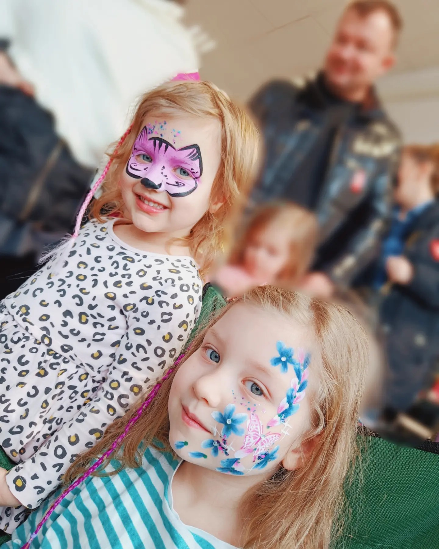 Dwie uśmiechnięte dziewczynki z pomalowanymi twarzami: jedna z motywem różowego kotka, druga z niebieskimi kwiatami i motylem.