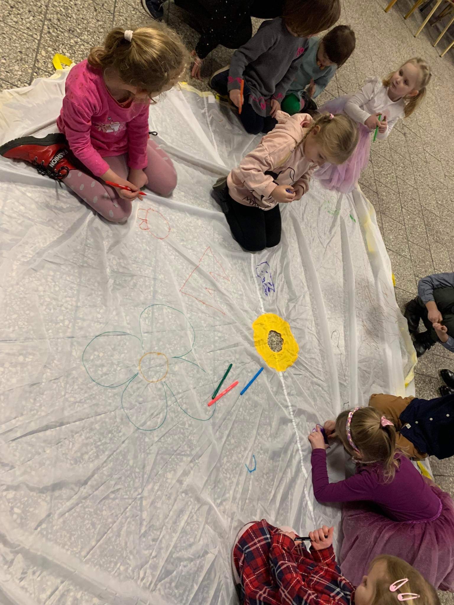 Grupa dzieci rysuje kolorowymi pisakami na rozłożonej, dużej płachcie materiału, tworząc kwiaty i inne wzory.