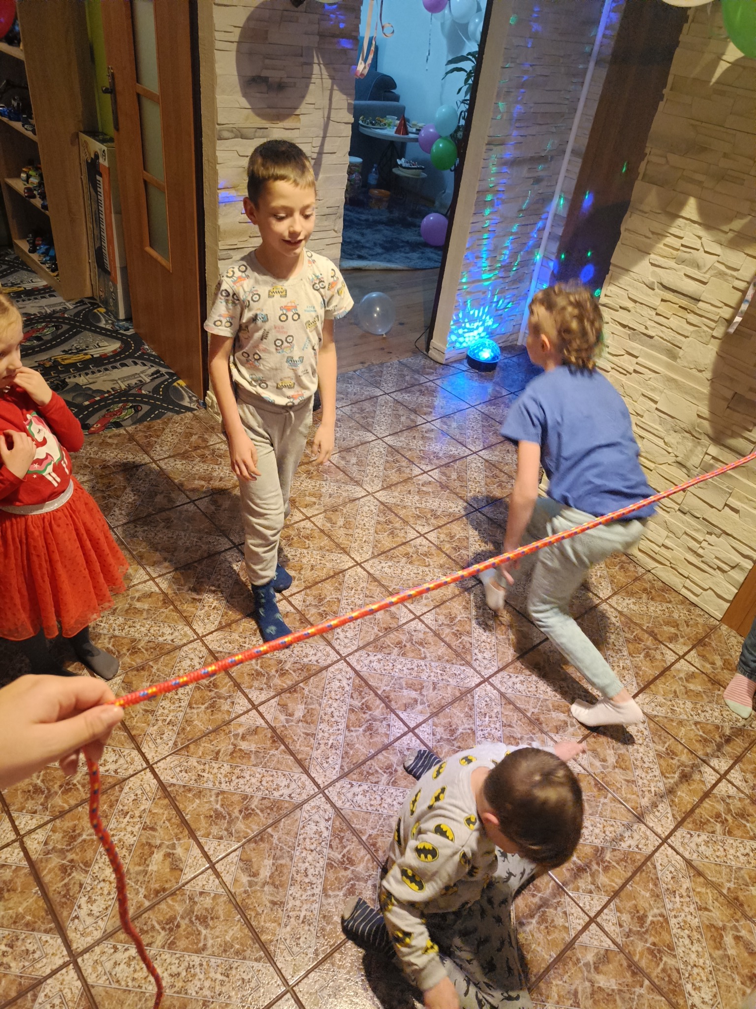 Dzieci bawią się w limbo na brązowej podłodze z płytek. Jeden chłopiec przechodzi pod kolorową liną, inny stoi obok, a dziewczynka w czerwonej sukience przygląda się.